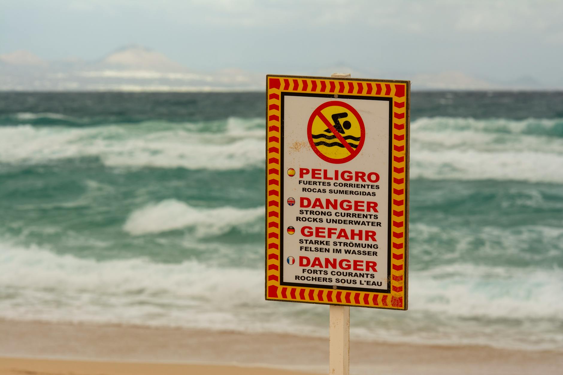 A beach warning sign cautioning about strong underwater currents in multiple languages.